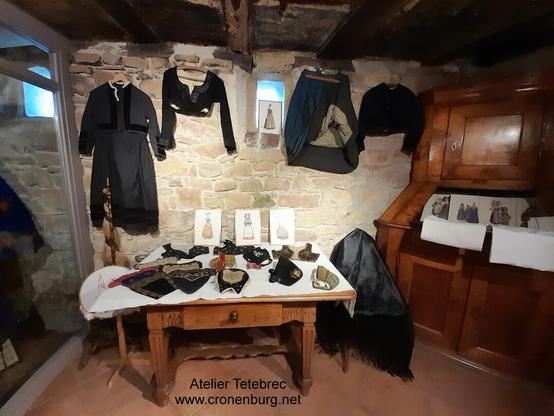 Bonnets and caps on a 19th-century table. Historic costumes hanging on the stone wall.