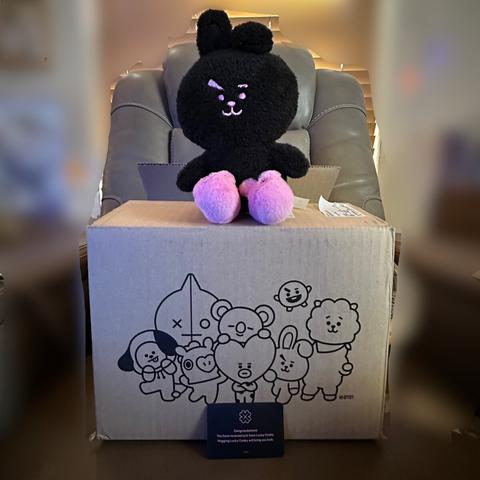 Black edition Lucky Cooky sitting on a Line Friends box with the other BT21 characters. Cooky is wearing pink bunny slippers. A small black card stands in front of the box and is weird in the tweet.