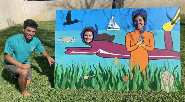 Our photo opp board, where 2 women (artists Makenzie and Natalie) are putting their heads through to look like they’re snorkeling in a seagrass meadow. Keith kneels next to the board, pointing at a shrimp he painted