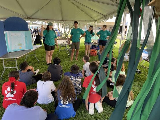 Group of elementary school students sitting on the lawn under a tent listening to a group of scientists introduce themselves