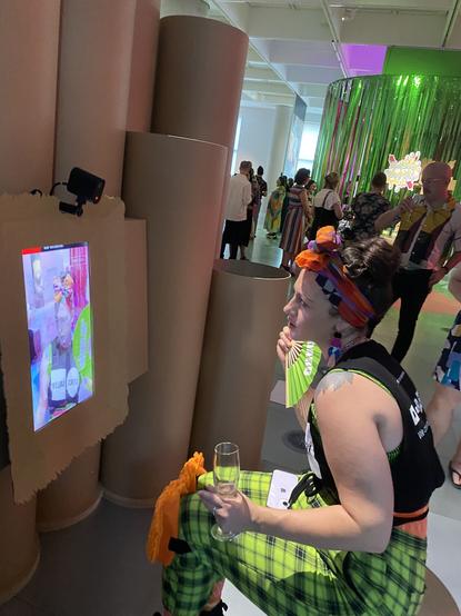 A person in a fabulous orange head wrap brown hair and neon clothes with a green fan interacting with self portrait, an augmented reality artwork using stablediffusion to create a mask and do a face swap.