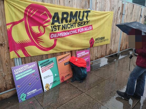 Ein Banner und Plakate an einem Holzbretterzaun, unter anderem mit dem Text "ARMUT IST NICHT SEXY - GEMEINWOHL FÜR ALLE!"