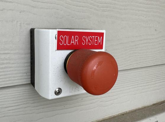A large red pushbutton attached to a building exterior with a small red engraved nameplate labeled “SOLAR SYSTEM”.