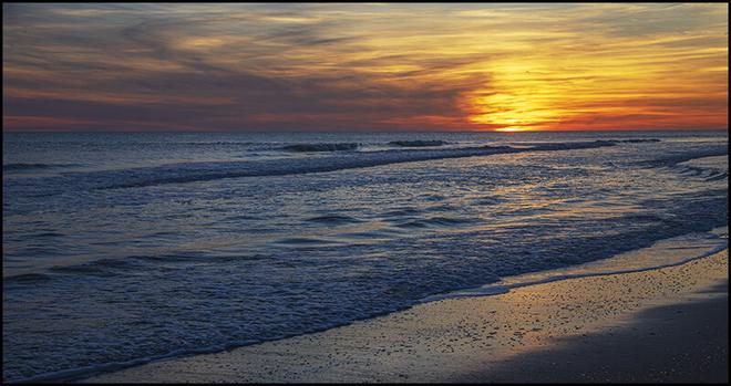 It is mid-February at Atlantic Beach NC. The weather is mild and the sky is a glow with hues or orange, yellow and red. Gentle waves wash up onto the sand. It is a beautiful sight to behold.
