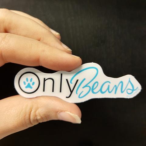 A hand holding a white sticker on a black background. The sticker is cut to the shape of text 'OnlyBeans', a parody of the former OnlyFans logo. 

The only text is black with a blue paw print in the O; the beans text is a swoopy blue cursive at a gentle angle
