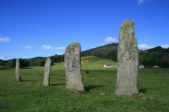 four standing stones ascending in height. They stand in a green field with a hill behind half covered in plantation. There are low white buildings at the foot of the hill. Blue sky, sunshine.