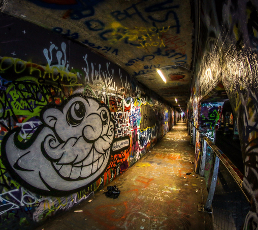 A car/pedestrian tunnel with every surface covered in colorful graffiti; darkness intermittently lit with overhead fluorescent lights; small figures of people walking in the distance