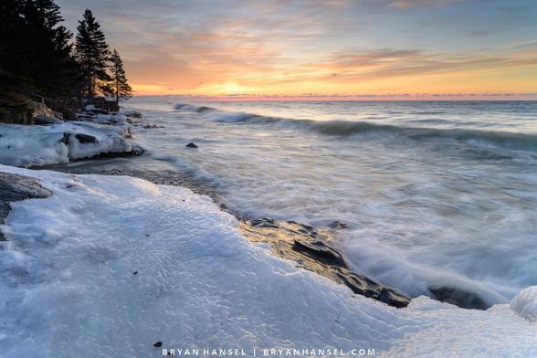 A blueish ice-covered shoreline leads to a large body of water on which a wave washed towards shore. The water is on the right of the image and the shore on the left. In the upper left corner of the photo, several fir stand upon a rocky point. The sunrise is turning the sky orange and yellow.