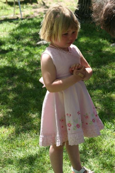 A blond little girl about 3 years old wearing a pink dress is standing on a sunny lawn, looking like she's been crying.