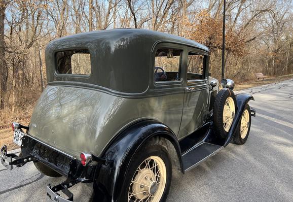 A green 1931 Ford Model A Victoria car, with yellow wheels on a tree-lined road.
