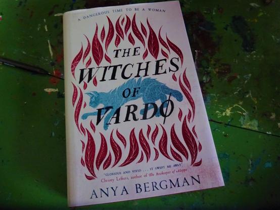 A book face up showing the cover - the text reads: The Witches of Vardo by Anya Bergman, A Dangerous Time to Be a Woman. The cover is white, with a print of red flames, and a blue lynx.