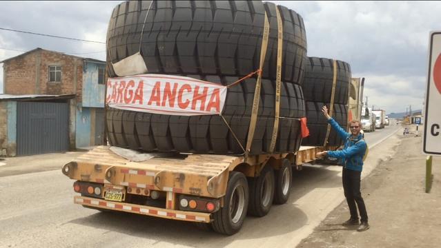A person gesturing toward the rear end of an articulated lorry, which is carrying four giant tyres. A warning sign strapped to the back reads "Carga Ancha" meaning "Wide load" in Spanish. The scene is dusty and overcast