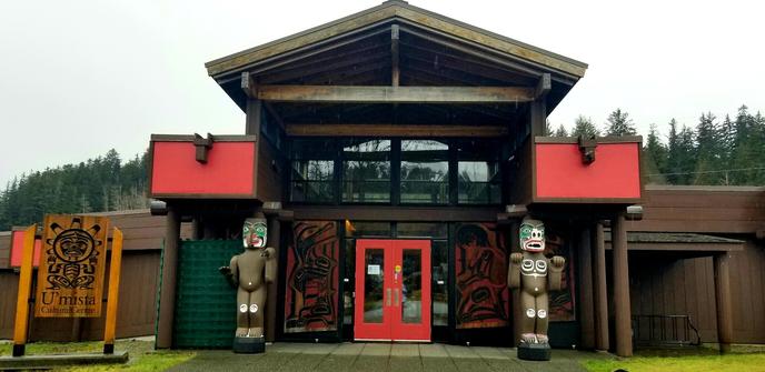 Front entrance to Umista Cultural Centre.  Two welcome totems flank both sides of walkway to doors.  Native art panels are placed on both sides of the entrance doors. The building sign is on the far left.