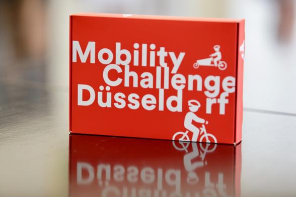 Eine Box, die auf einem Tisch mit spiegelnder Oberfläche steht. Die Box ist in roter Farbe gehalten. Auf dem Deckel steht in weißer Schrift: "Mobility Challenge Düsseldorf". Den Deckel zieren zudem stilistische Darstellungen eines Radfahrers und den Fahrer eines Lastenrades.