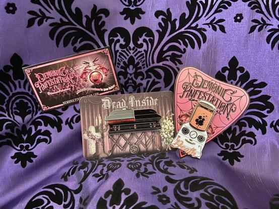 A purple damask background featuring two enamel pins on card stock backings. A business card reads “Demonic Pinfestation.” The pins are shaped as an embalming machine and a casket.