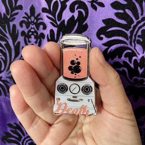 A purple damask background with a Caucasian hand holding a small enamel pin. It’s in the shape of an embalming machine with pink fluid in the tank and a pink hose shaped into the word “death.”