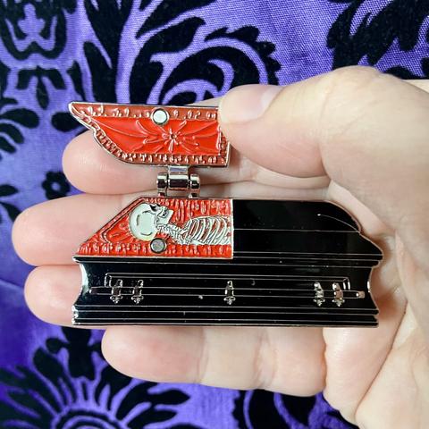 A purple damask background with a Caucasian hand holding an enamel pin shaped like a black casket. Half of the lid is open to display a red interior and a white skeleton.