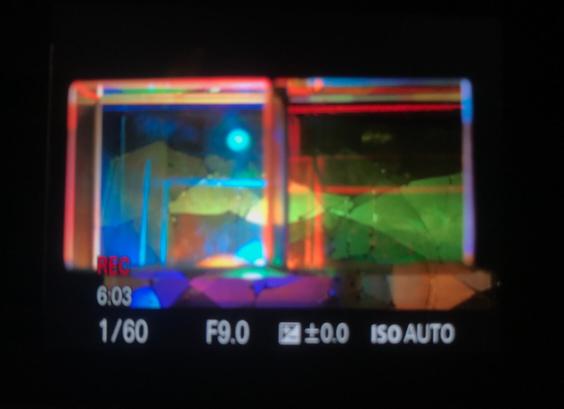 Two glass cubic prisms, saturated with the full spectrum of colour stand in front of a cracked screen whose shards resemble a mountainous landscape. A reflected pale blue light source hangs moon-like above them. All of this is seen soft focus on the back screen of a camera showing that is is recording, with various numbers telling the shutter speed, aperture and so on.