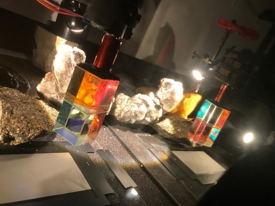 A surface, strongly lit with a single light sourec and cluttered with various rocks. Two cubic prisms glued together hang in the foreground. The whole scene is doubled to the right by a mirror's reflection.