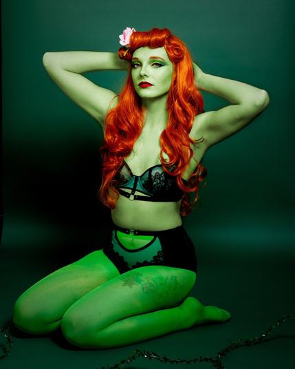 A green skinned woman with long red hair, Poison Ivy, kneels in front of a green background whilst wearing green lingerie