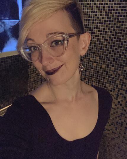 It’s a bathroom selfie! Sonja is blonde and wearing sparkly translucent cateye glasses; their eye makeup is dark but understated and their lipstick is black. They’re wearing a long sleeved dark purple bodycon dress with a low horseshoe neckline. They’re standing against the tiled background of a nice looking bathroom; the corner of a lightbox featuring xray panels can be seen behind them. 

It’s a soft goth vibe and Sonja is cute.