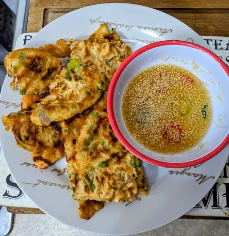 Torn Korean pancakes next to a small bowl of dip, which is topped with sesame seeds, on a white plate