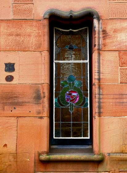 A narrow stained-glass window in a red sandstone building with a curved lintel above it.