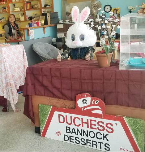 Me & Alert Bay's Duchess of bannock.  I'm at table with her small business sign, wearing a big rabbit's head.  Duchess is standing behind counter, in background.