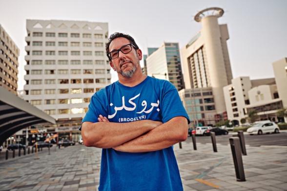 A white guy with dark hair, glasses and a beard, with arms crossed over a blue t-shirt reading “Brooklyn” but written in Arabic, standing in front of modernist architecture in downtown Abu Dhabi