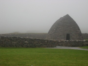 Gallarus Oratory - entirely stone church shaped like an upturned ship. One small door surrounded by bare stone wall. A lawn of green grass to the fore. The background is shrouded in fog.