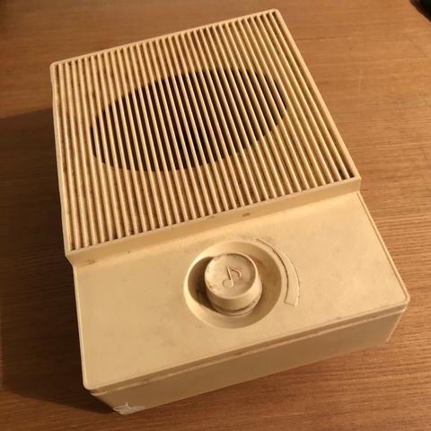 Closeup of the Ukrainian radio as it appears now. The top two thirds of the all-plastic device is devoted to a single mono speaker. Beneath the speaker, a single circular dial embossed with a musical eight note sits in a recessed indentation in the body of the radio. Alongside the indentation, a directional arrow points clockwise indicating the volume will increase when the dial is turned to the right. The radio needs a good cleaning, which we should finally get around to doing this year.