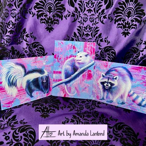 A purple damask background featuring three 5x7 art prints. Each has a matching abstract background of pinks and teals  one picture is a realistic painting of a skunk, one is an opossum, and one is a raccoon.