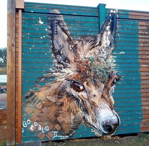 Installation. An art installation on a high wooden fence. The image of a small donkey's head has been assembled from garbage. Cardboard parts as neck, wires as mane and many other materials have been combined with painted parts to create a wonderful little mural.
