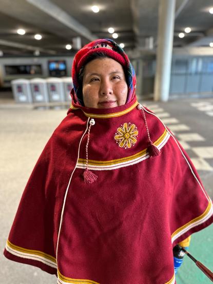 Maria is clad in a red luhkka with yellow sun ornament on it. They’re looking solemnly ahead.