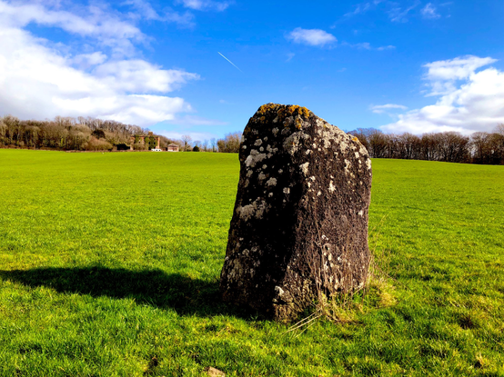 a chunky standongstone in a green field on a sunny day. In the distance you can just see a partially ruined mansion house