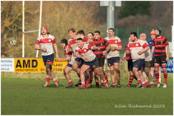 Colour photograph of two male rugby teams following a ball. One team wearing red and black, the other wearing red and white