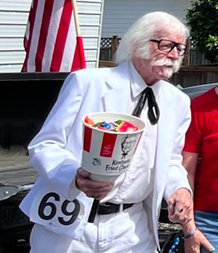 zoomed in shot of one of the participants, who is inexplicably dressed like colonel sanders (KFC) and carrying a sign that reads "69" as well as a KFC bucket filled with something that's hard to see.