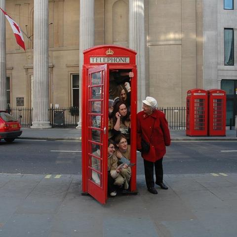 A group of people crammed into a red London phone booth as an older woman in a red coat looks on