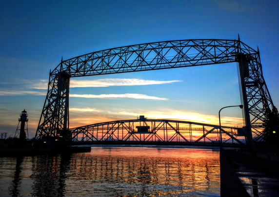 Landscape photograph of a bridge with a sunset behind it -- the sky above the orange/yellow sunset is very blue with just a few wispy clouds near the horizon.  The bridge is constructed of metal members that form triangular shapes, so the sunset shows through most of the bridge structure.