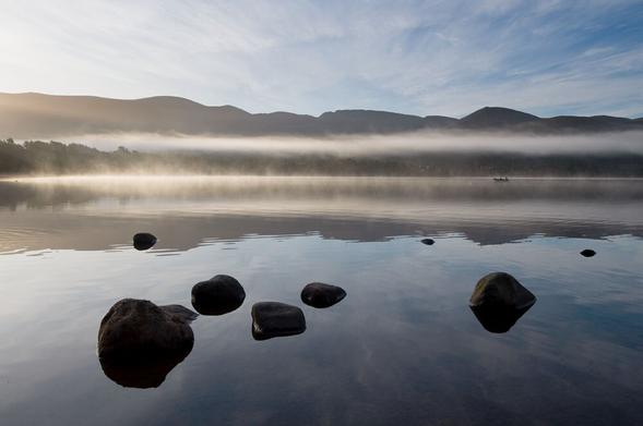 A landscape photograph of Loch Morlich on a misty morning. In the distance is a band of hills with a narrow bank of cloud hanging halfway up the hillside and across the whole of the view. In the middle ground light mist is rising from the loch. A small rowing boat is in the rising mist on the right. The band of cloud and the hills are reflected in the almost still water. Prominent for foreground interest is a number of rounded stones in the loch. Light high cloud/blue skies radiate from the centre upwards and are mildly reflected in the loch