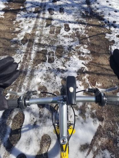 A photograph shows the perspective looking down over a pair of handlebars belonging to a yellow bike.  There is snow on the ground below, along with footprints, paw prints, and tire tracks in the snow.
