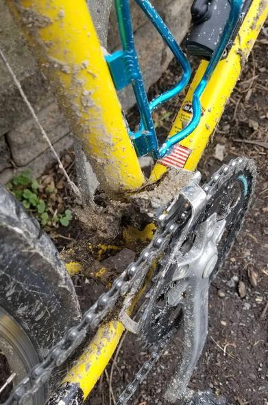 A yellow bicycle's front chain ring is caked in mud from the back tire.  Muddy ground is beneath.