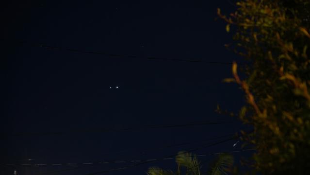 Dark blue sky, blurry bushes off to the right. In the middle of the sky are two bright spots right next to each other, the right one noticeably brighter.
