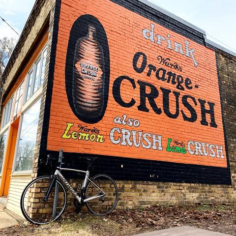 Large vintage ad painted on side of a brick building for Orange Crush soda. The wall is mostly orange with an image of a bottle of soda and the words, Drink Ward’s Orange Crush also Ward’s Lemon Crush and Ward’s Lime Crush. Below the sign s silver bicycle leans against the building.