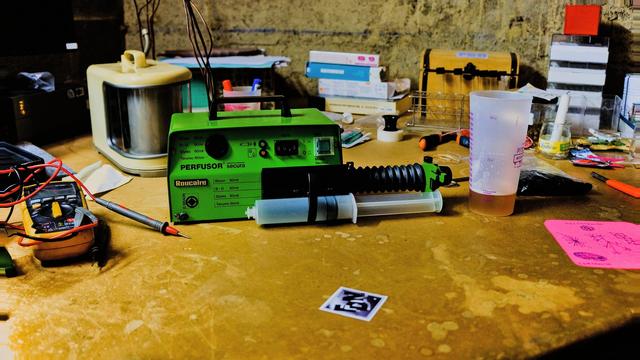 Photographie d'un appareil dit pousse-seringue de couleur verte sur un plan de travail en bois jaune-vert avec une seringue de 50 ml ajustée dans l'appareil