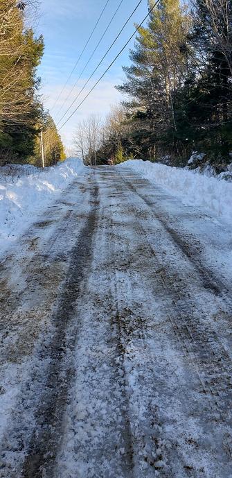 Plowed icy dirt road looking uphill. High snowbanks on either side which is lined with tall trees. The sky is blue with a few small wispy clouds.
