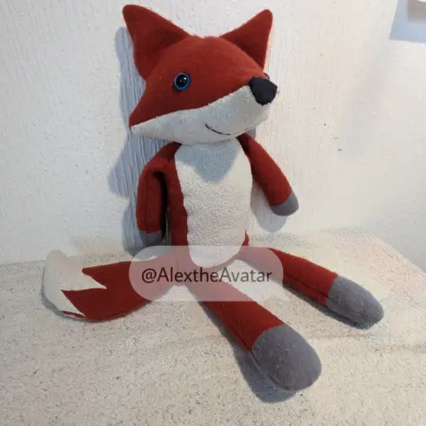 Ein kleiner rot-weiser Plüschfuchs mit grauem Pfoten und blauen Augen blickt fröhlich in die Kamera.