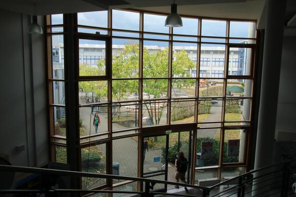Aus erhöhter Sicht, eine Treppe aufwärts, schaut die Kamera herunter auf einen Eingangsbereich. Neben der Glastür und, noch viel mehr darüber, sind viele große Fensterscheiben, nur durch Rahmen unterbrochen. Draußen ist ein großer Hof, auf dem Bierbänke und -tische aufgebaut wurden. Der Himmel ist etwas dunkler bedeckt.