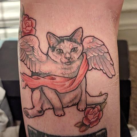 Tattoo of a fat, derpy cat sitting with rear legs splayed, cherub wings, and a pink sash. It is framed with two roses on either corner. The wings have touches of pink and blue, making them almost pearlescent.