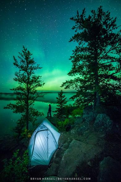 Green Northern lights reflect over a lake. A tent and person are in the foreground.